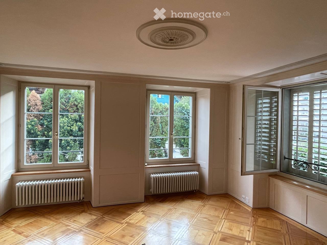 Empty room with wooden flooring, large windows, radiators, and a decorative ceiling light fixture.