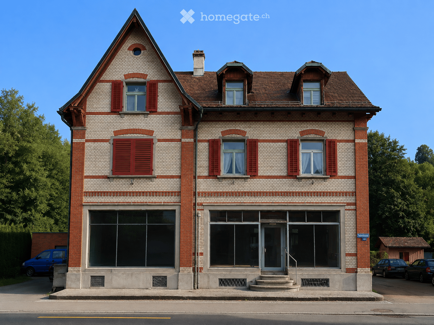 Two-story brick building with red shutters, large windows, and a triangular roof. The ground floor has large glass windows and a central entrance with steps.