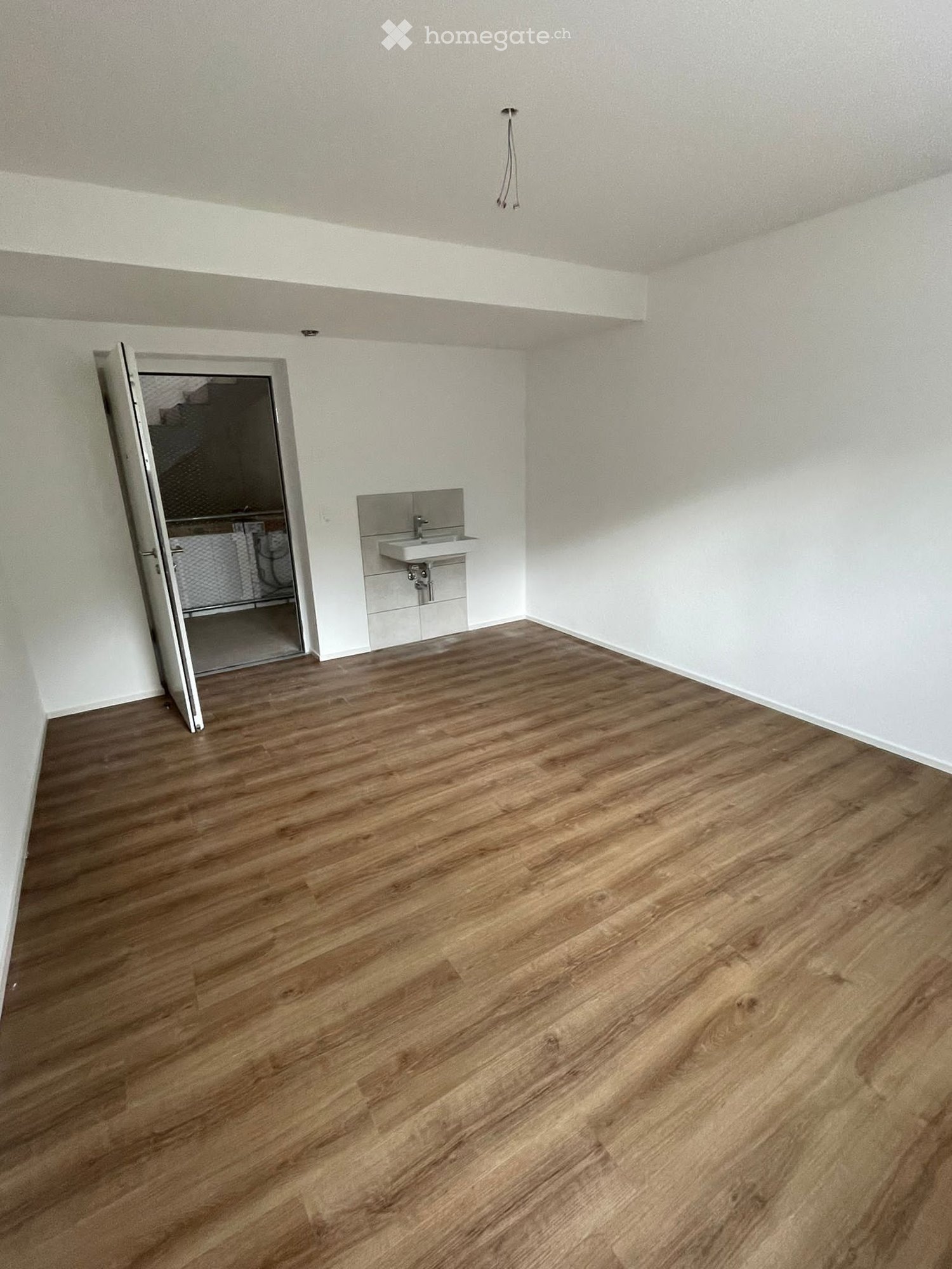 Empty room with wooden flooring, white walls, and a small sink in the corner. An open door leads to another space.