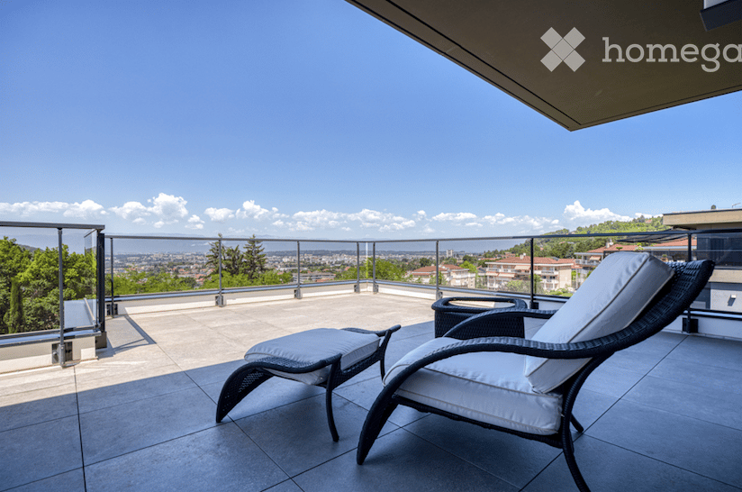 A modern balcony with a panoramic city view, featuring a wicker lounge chair and matching footrest, a small black side table, and a sleek metal railing.
