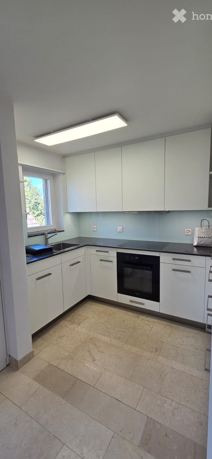 Modern kitchen with white cabinets, a black countertop, a sink, and an oven. The space features tiled flooring and a large window letting in natural light.