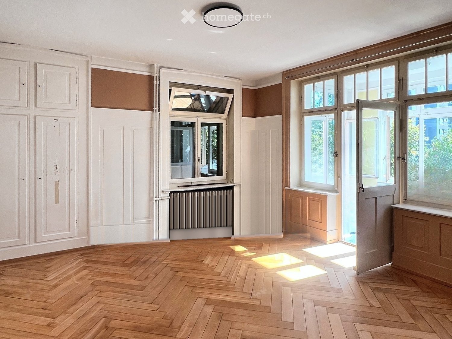 Empty room with wooden herringbone flooring, white walls, large windows, and a radiator. No furniture or personal items present.