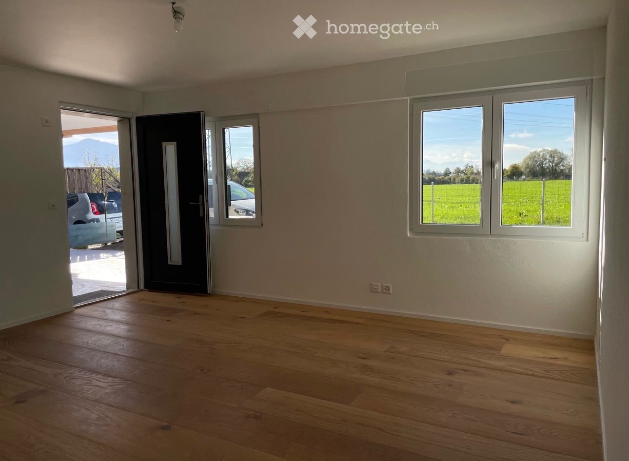 Empty room with wooden flooring, large windows, and a door leading outside. No furniture or equipment present.