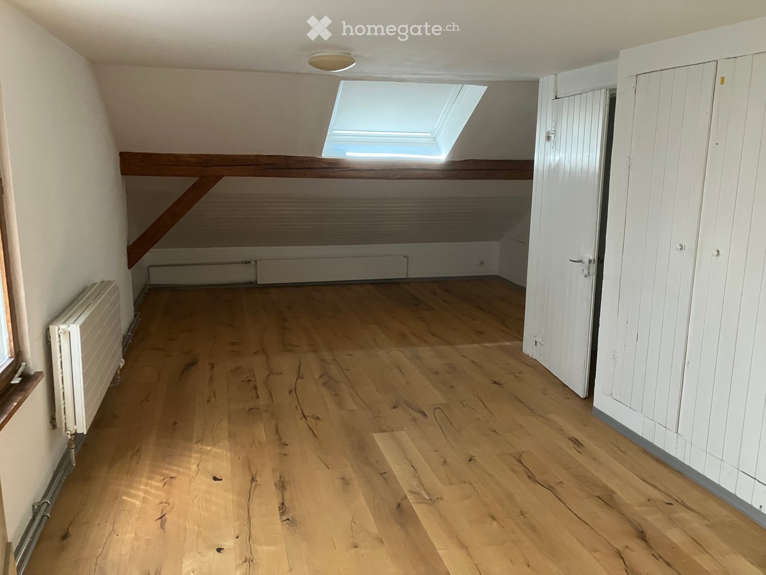 Empty attic room with wooden flooring, white walls, built-in storage, and a skylight window providing natural light.
