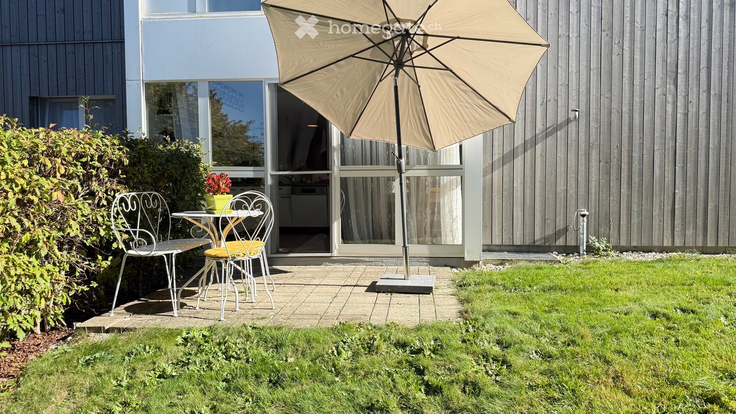 A small patio area with a table, two chairs, and an umbrella, adjacent to a modern house with large windows and a view of the interior.