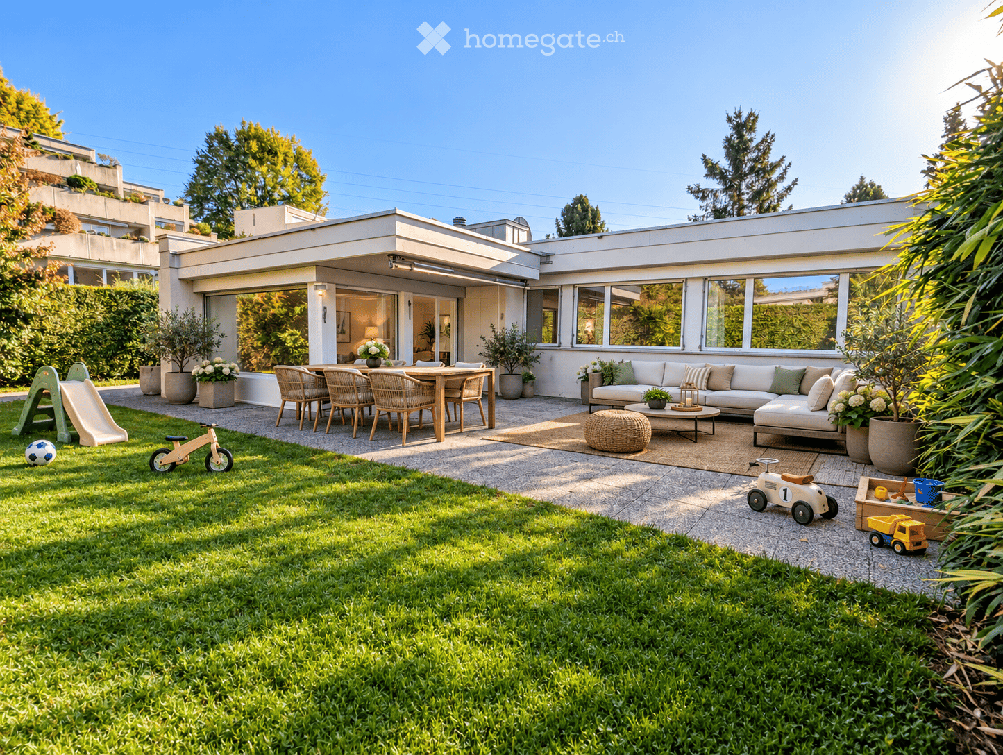 A modern outdoor space with a well-maintained lawn, a patio area with a sofa, coffee table, ottoman, and dining set, surrounded by potted plants and trees. Includes children's toys like a slide, bicycle, toy car, and truck.