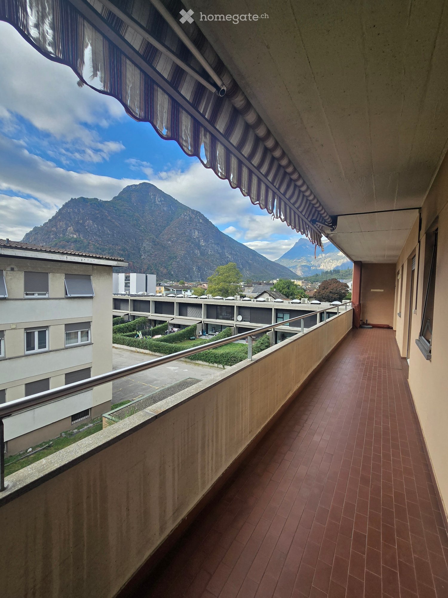 Balcony with red-tiled flooring, metal railing, and a striped awning, overlooking a scenic mountain view and surrounding buildings.