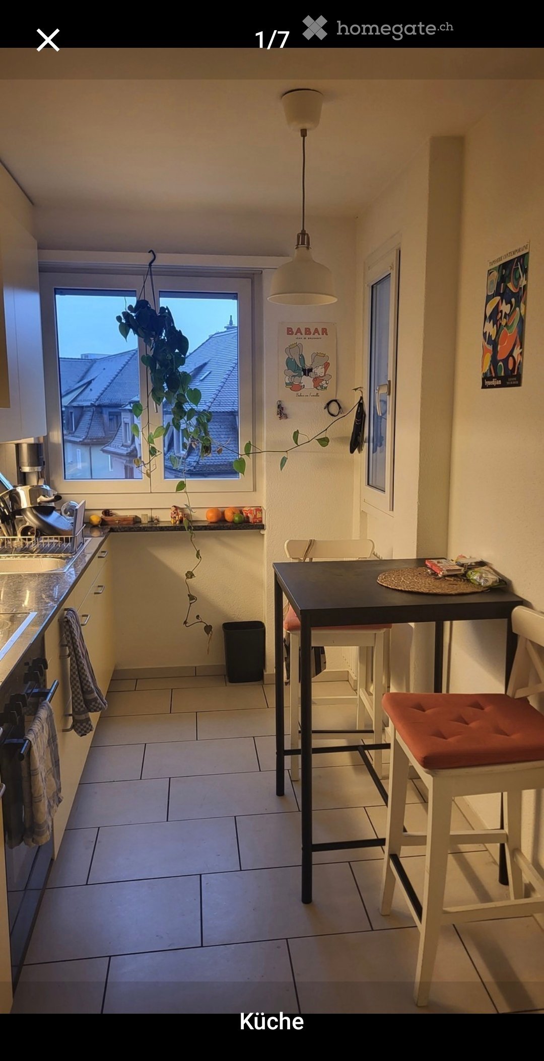 A compact kitchen with a sink, stove, and a small dining area featuring a table and chairs. The space includes a pendant light, a hanging plant, and various kitchen items on the counter.