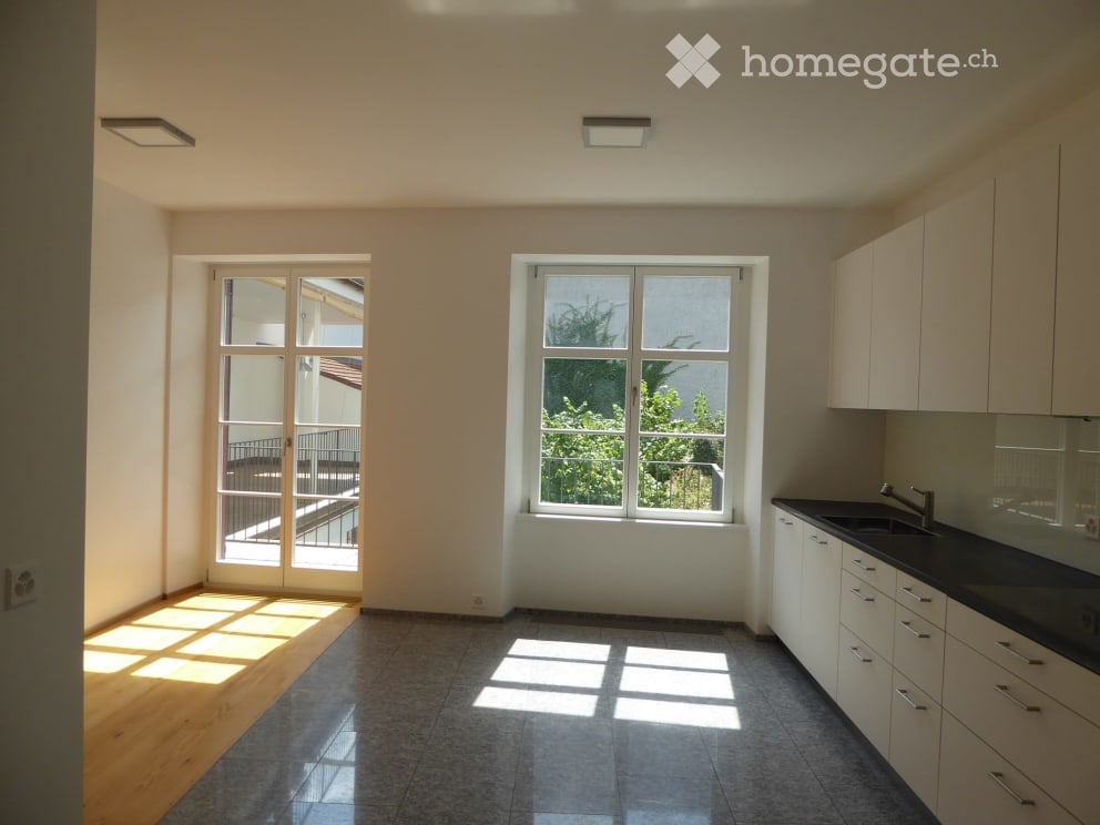 Empty kitchen with white cabinets, black countertop, and large windows, including a sliding door leading to a balcony.