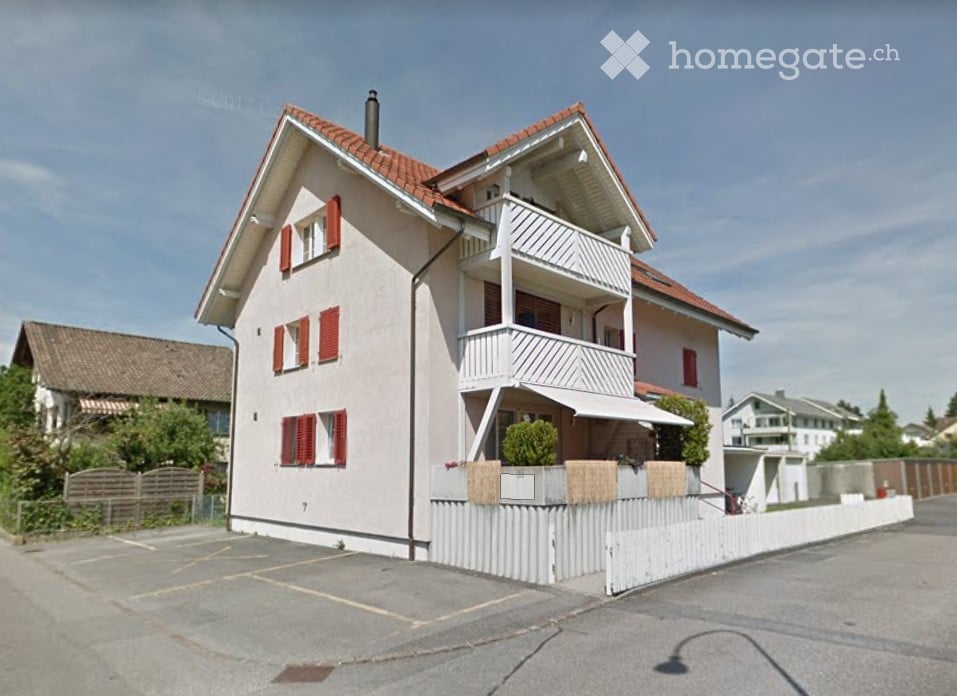 Two-story house with red shutters, white balcony railings, and a parking area.