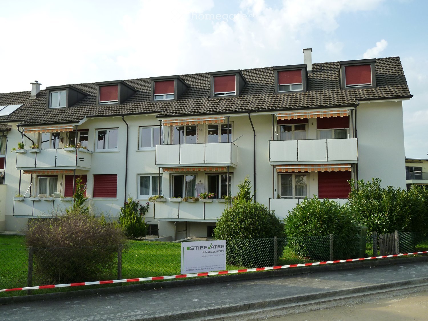 A two-story residential house with white walls, multiple windows, balconies, and a tiled roof. The house is surrounded by greenery and a fence.