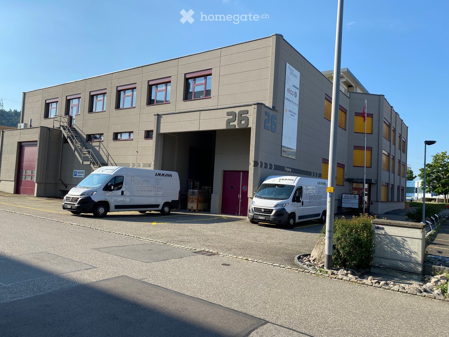 Two-story industrial building with large garage doors, two white vans parked in front, and signage for 'AMMANN Raum gestalten'.