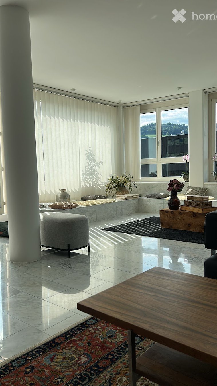 Bright living room with large windows, white walls, polished tile floor, and a mix of modern and traditional furniture including a sofa, coffee table, ottoman, and decorative vases.