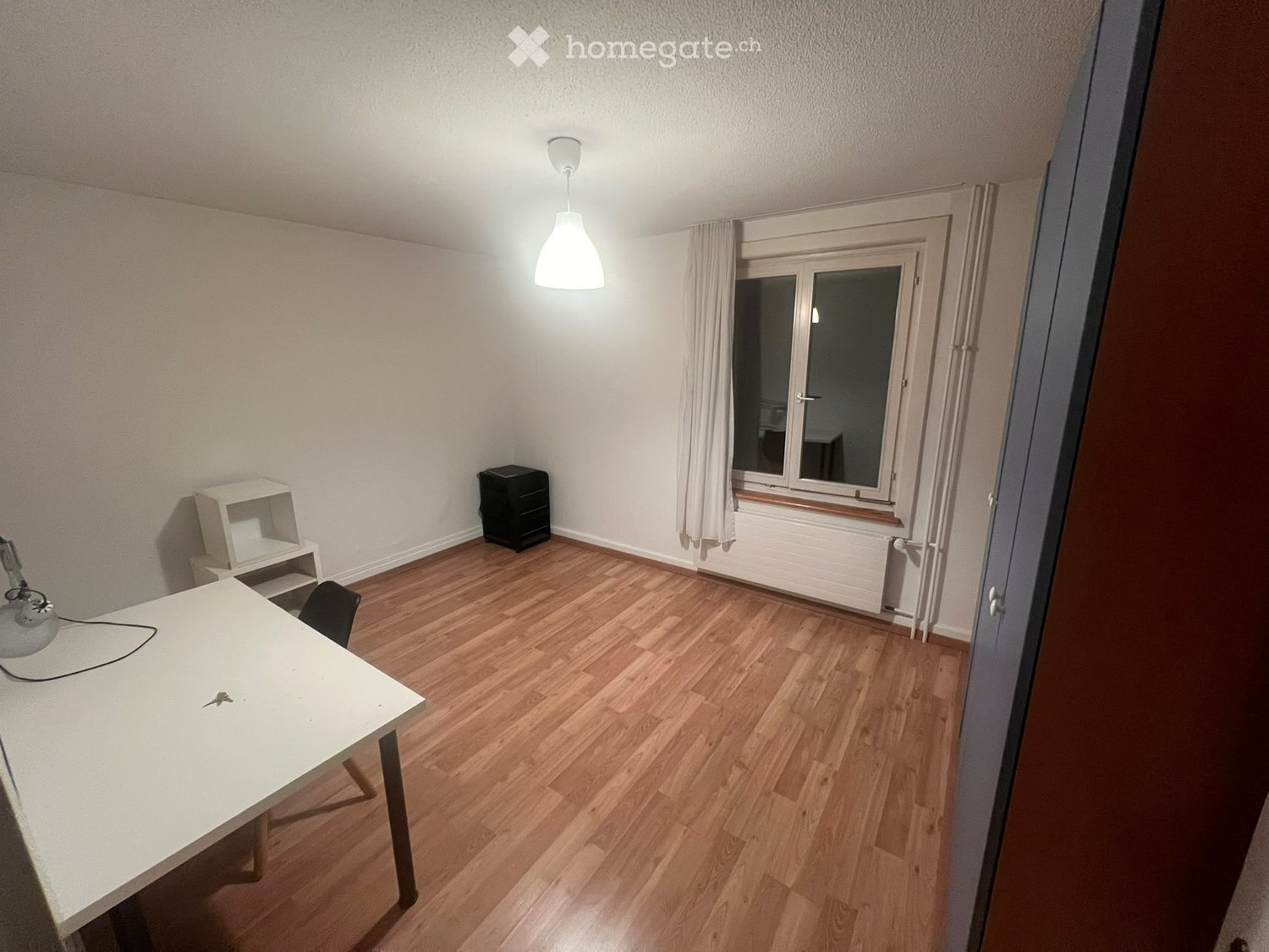 A small, unfurnished room with wooden flooring, a white table, a chair, a small shelf, and a lamp.