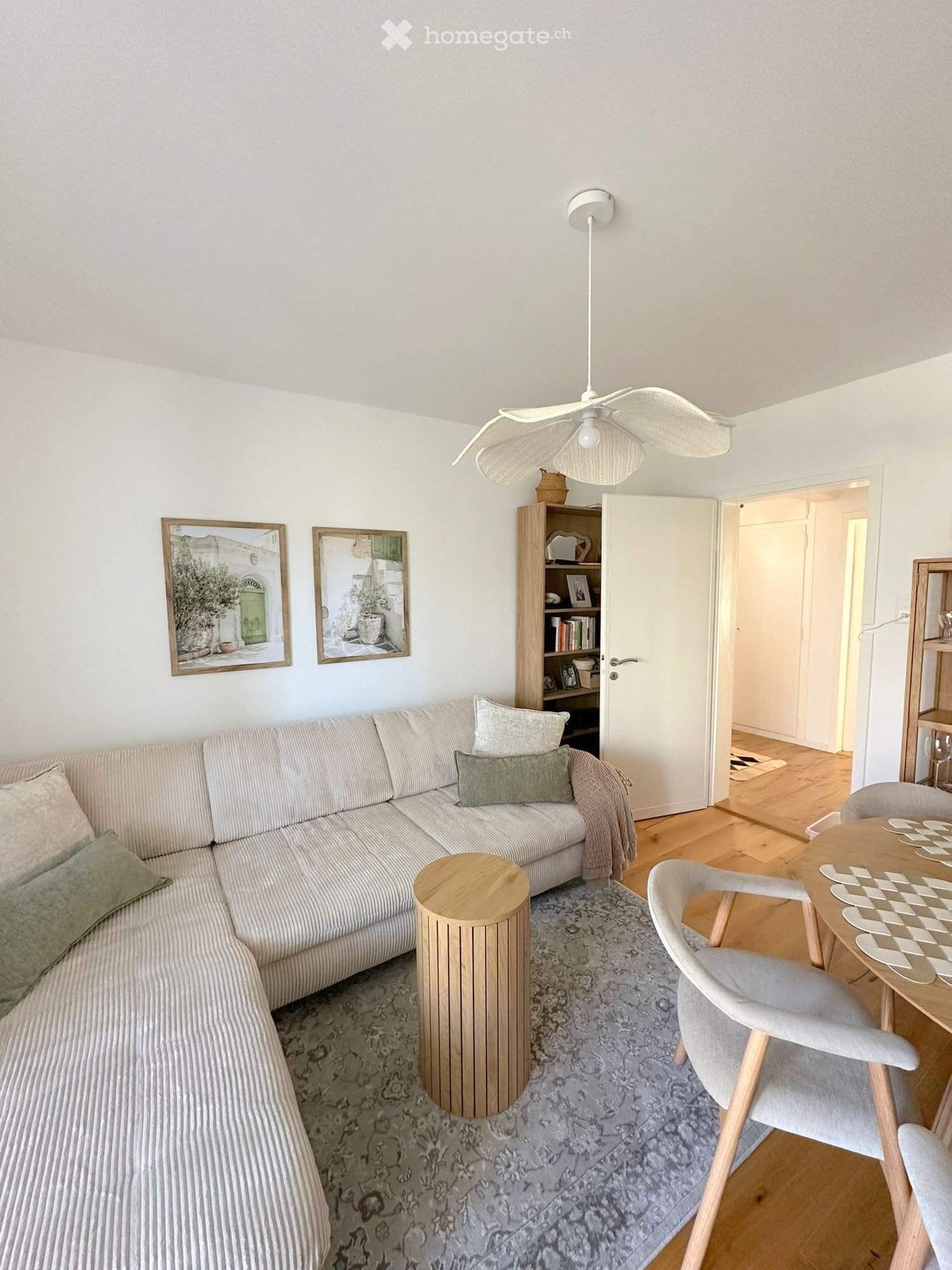 A modern living room with a light-colored sectional sofa, wooden coffee table, dining table with chairs, bookshelf, and decorative picture frames on the wall.