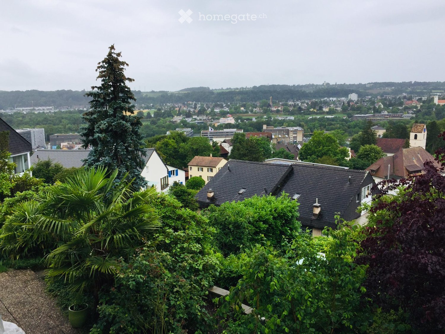 A lush garden with various plants and trees, overlooking a town with houses, buildings, and a church tower in the distance.