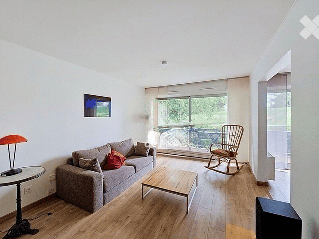 Bright living room with wooden flooring, large windows, and a balcony. Features a grey sofa with throw pillows, a coffee table, a rocking chair, a table lamp, and a television.