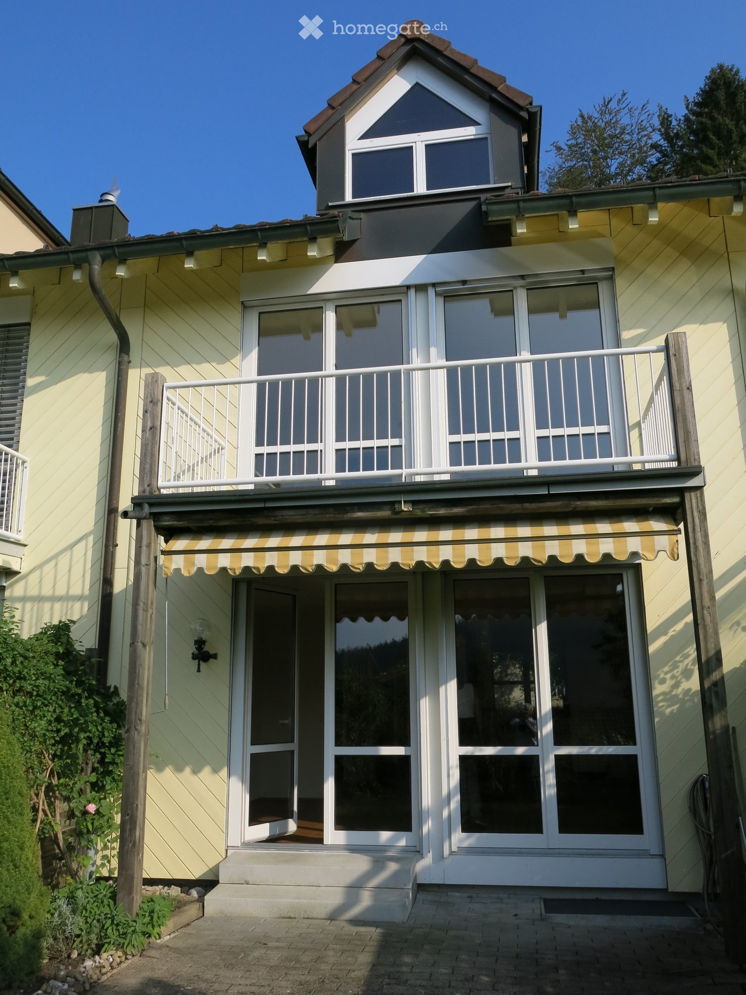 Two-story house with yellow siding, white trim, and a balcony with striped awning. Features large windows and a raised ground floor entrance.