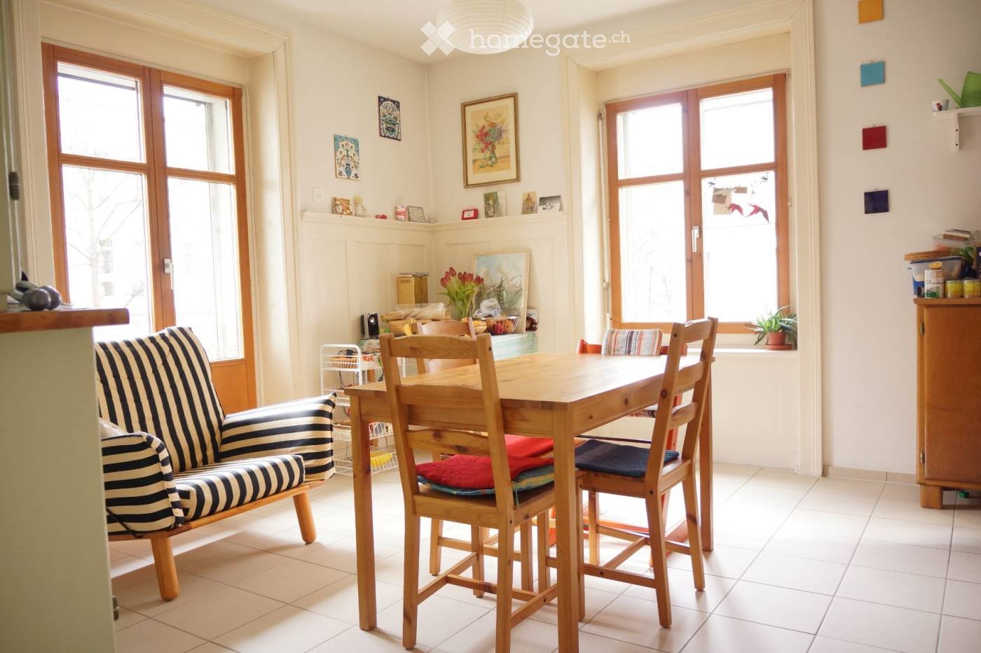 Brightly lit room with large windows, a wooden dining table with chairs, a striped sofa, and decorative items on shelves.