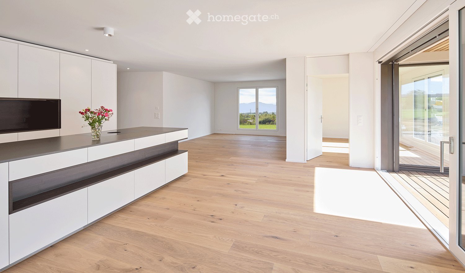 Modern kitchen with white cabinetry, wooden flooring, and a large island countertop. A vase with flowers is placed on the island, and there is a large window offering a scenic view.