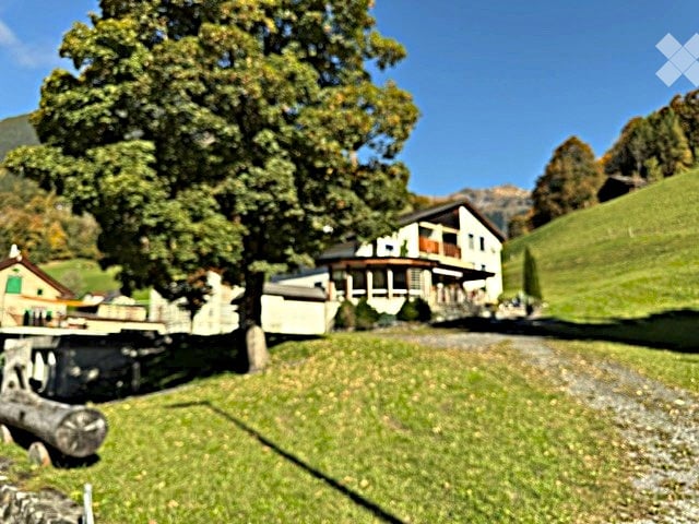 A picturesque rural setting with a large, traditional white house featuring balconies and a stone foundation, surrounded by lush green grass, trees, and rolling hills under a clear blue sky.