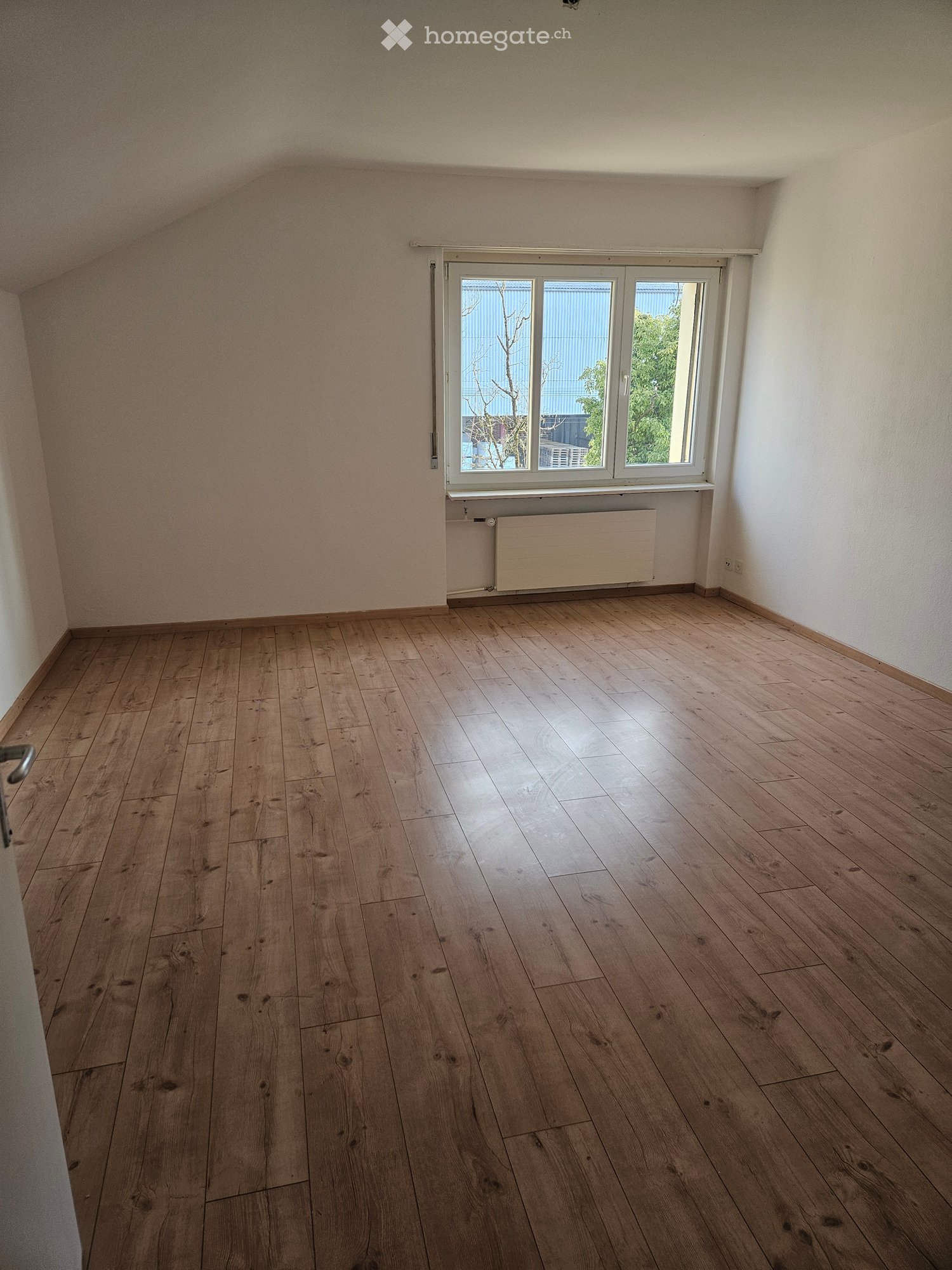 Empty room with wooden flooring, white walls, and a large window with a radiator below it.
