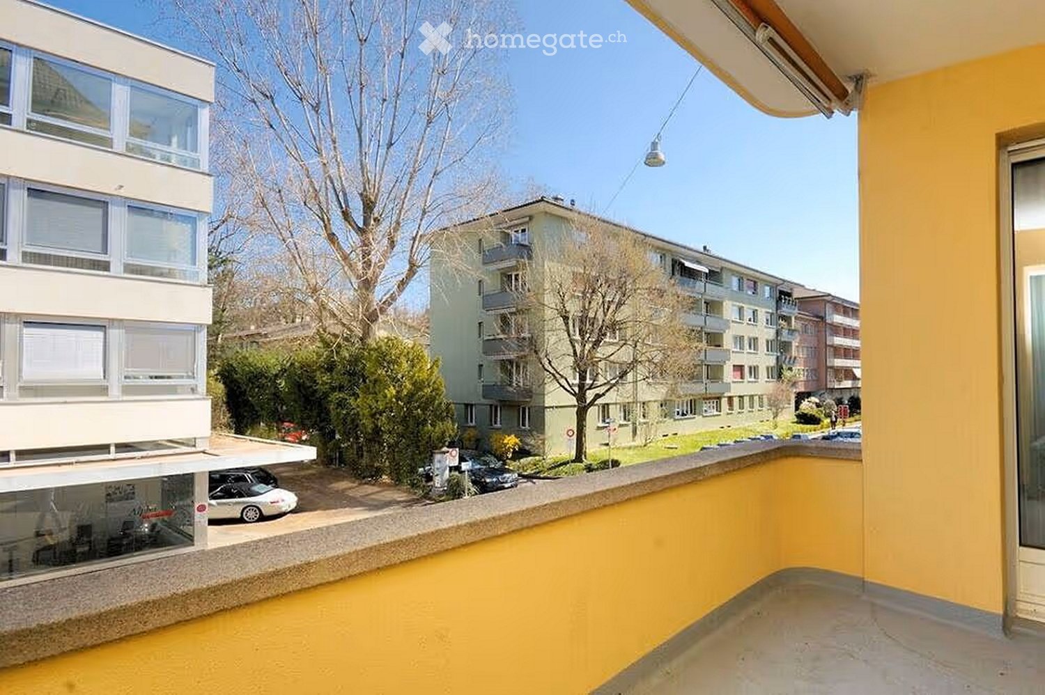 Balcony with yellow walls, overlooking a residential area with buildings, trees, and parked cars.