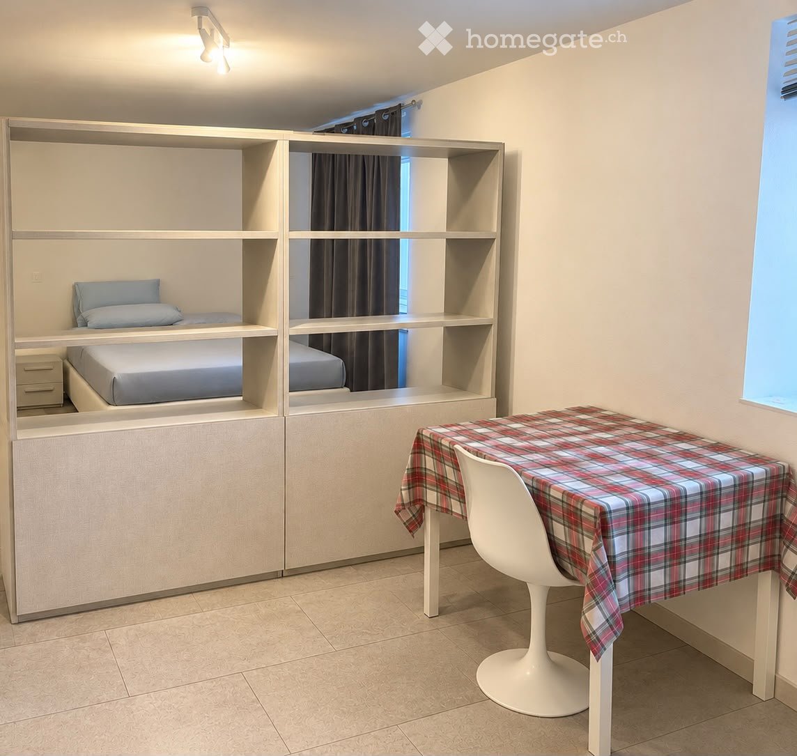 A compact, modern bedroom with built-in bunk beds, a small table with a plaid tablecloth, and a white chair. The room features light-colored walls, tiled flooring, and built-in shelving units.