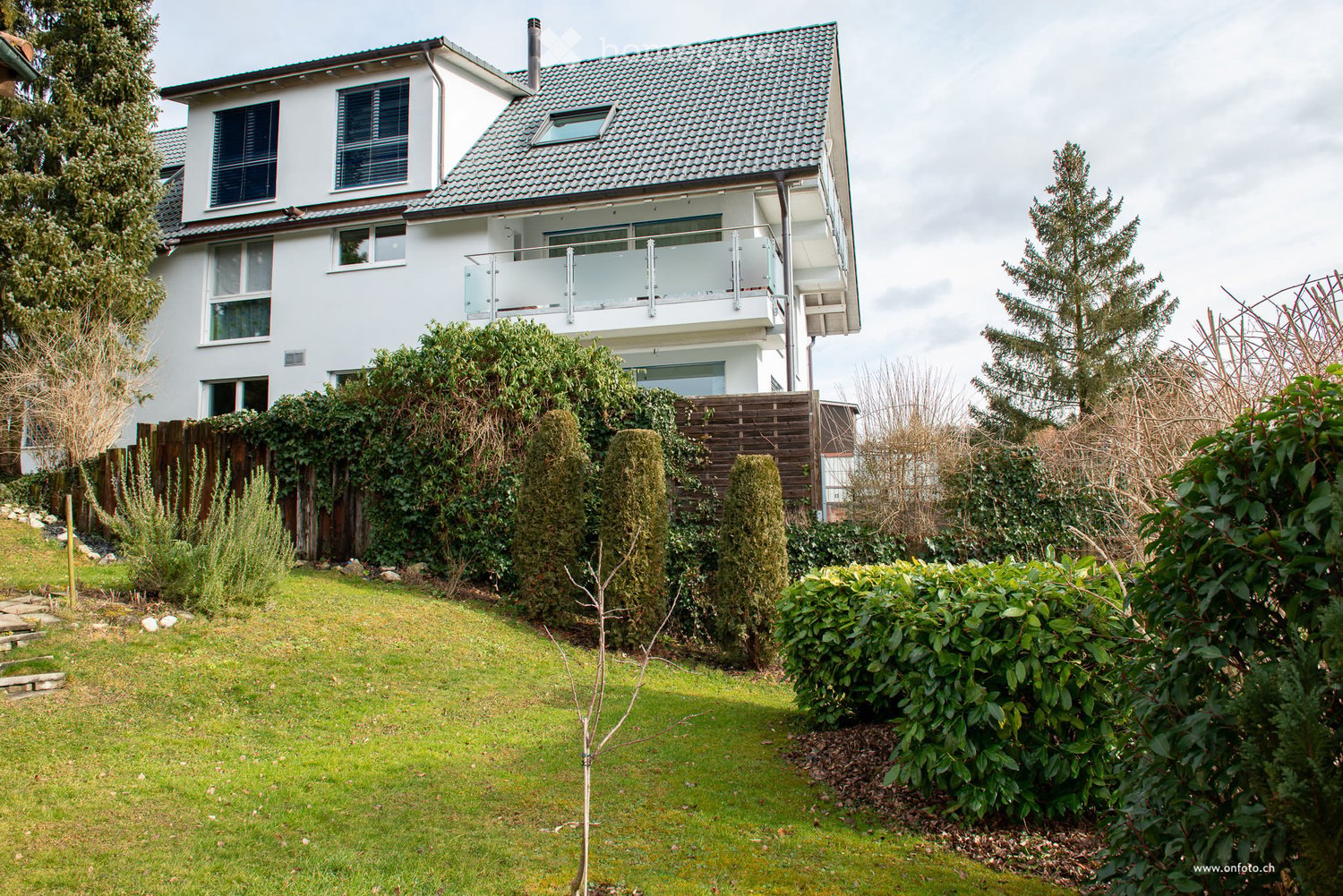 A two-story white house with a modern design, featuring a balcony on the upper floor, surrounded by a well-maintained garden with various shrubs and trees.