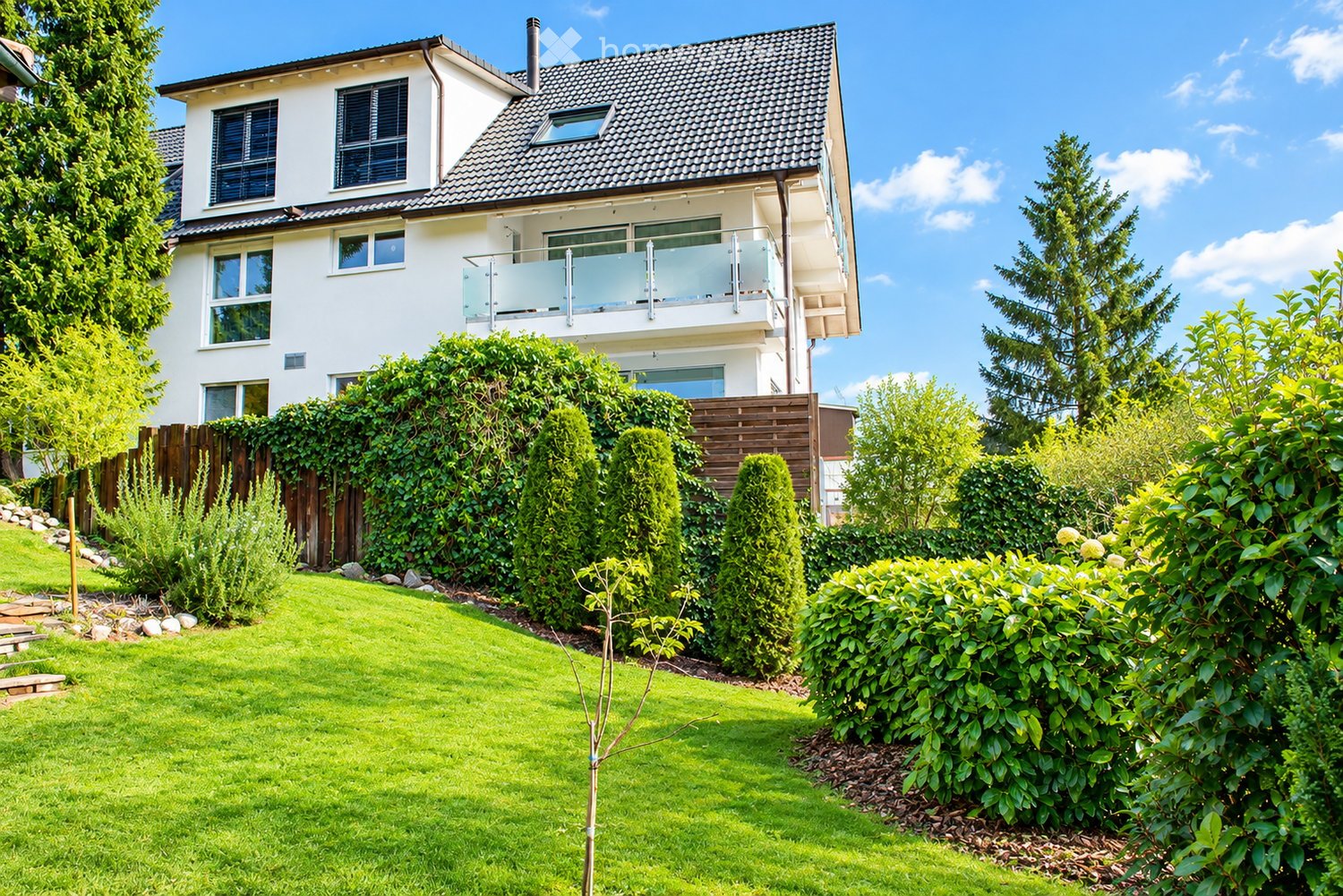 Two-story house with white exterior, multiple windows, and a balcony on the upper level. The house is surrounded by a well-maintained garden with green lawn, shrubs, and trees.