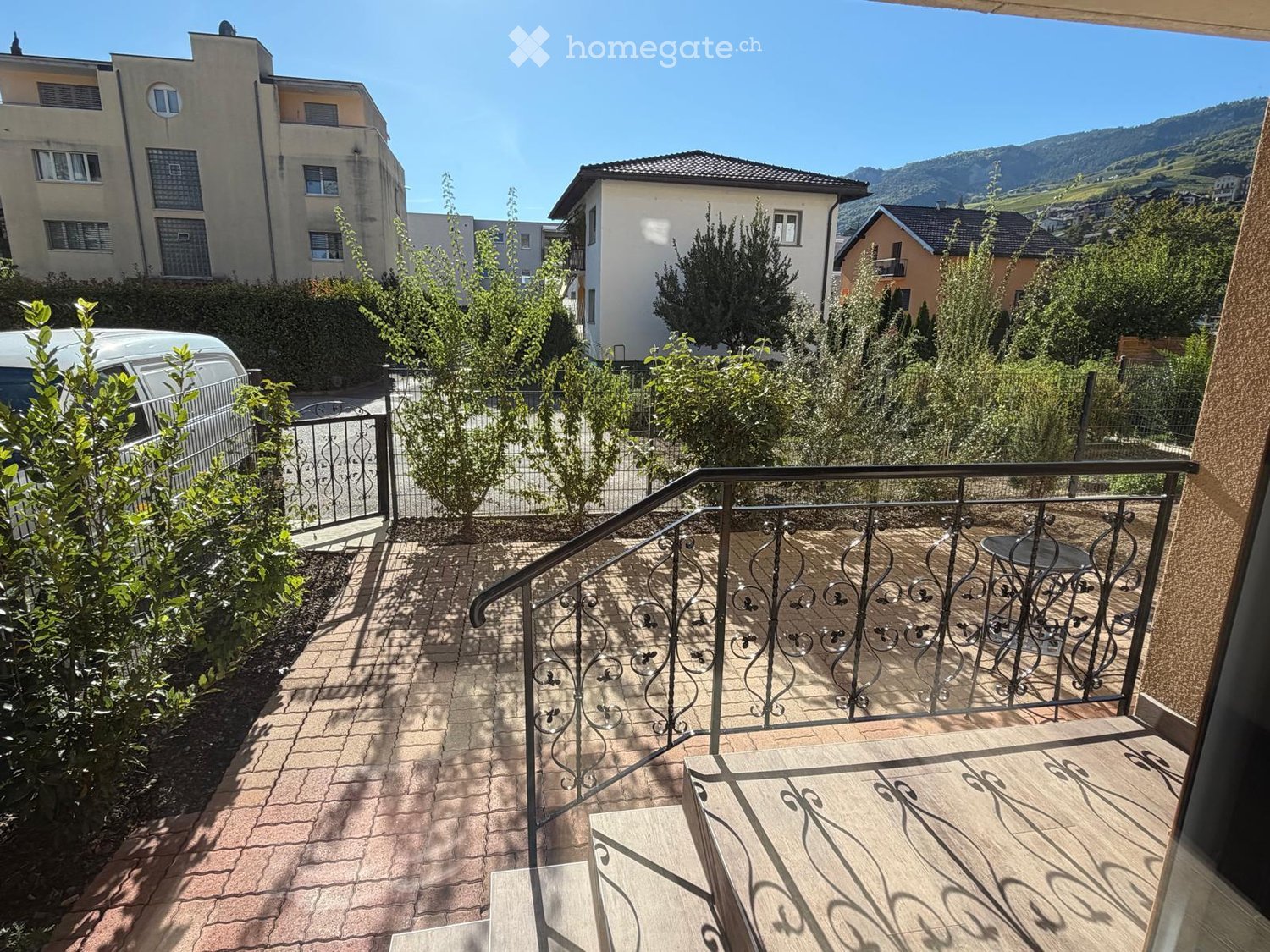 Balcony with wooden decking, metal railing, small table and chair, overlooking a residential area with trees and mountains.