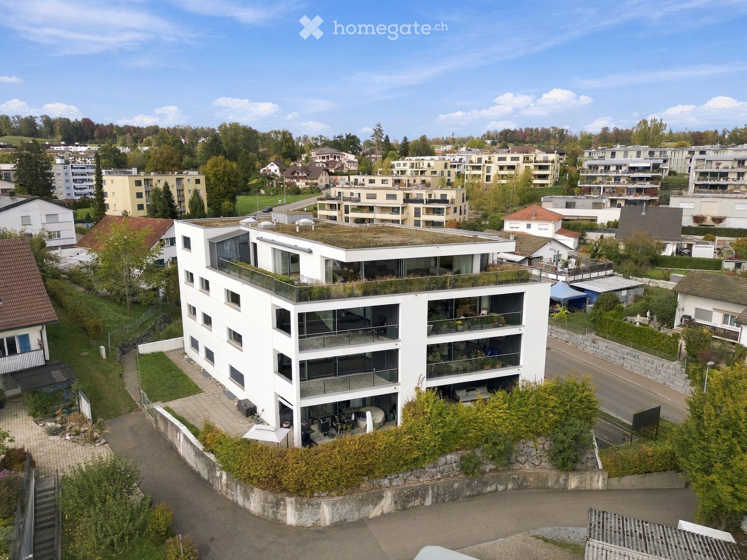 Modern white multi-story building with large windows, balconies, and a green rooftop, situated in a suburban neighborhood with other residential structures and greenery.
