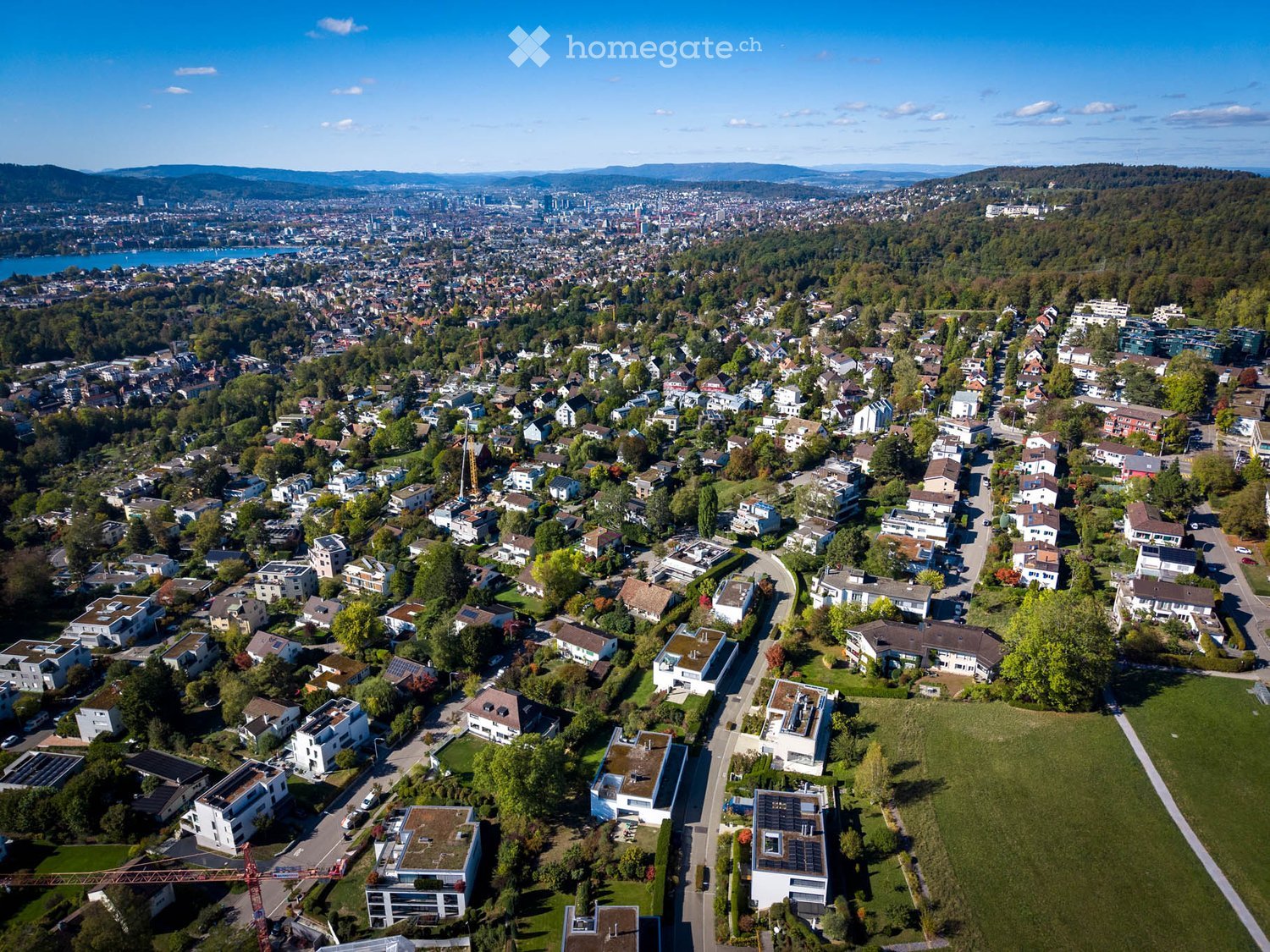 Aerial view of a suburban neighborhood with numerous houses, green spaces, and a distant cityscape with a lake and mountains in the background.