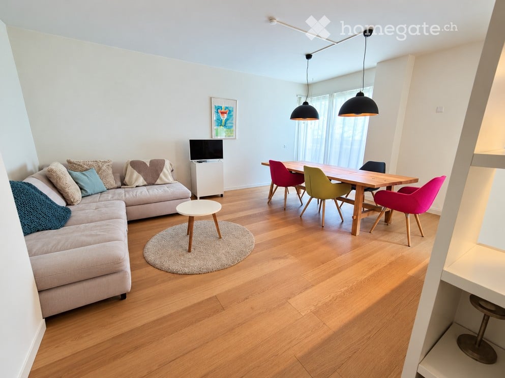 Open-plan living and dining area with light wood flooring, a grey sectional sofa with colorful cushions, a small round coffee table, a dining table with colorful chairs, and a television on a stand.