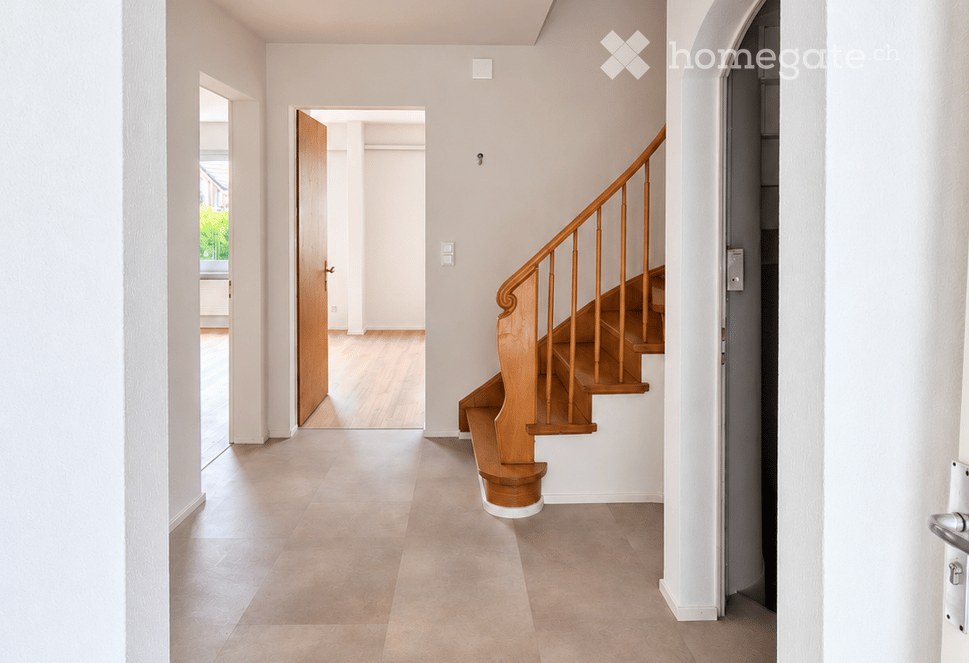 Empty hallway with a wooden staircase leading upwards, light-colored walls, and a tiled floor. An open door leads to another room.