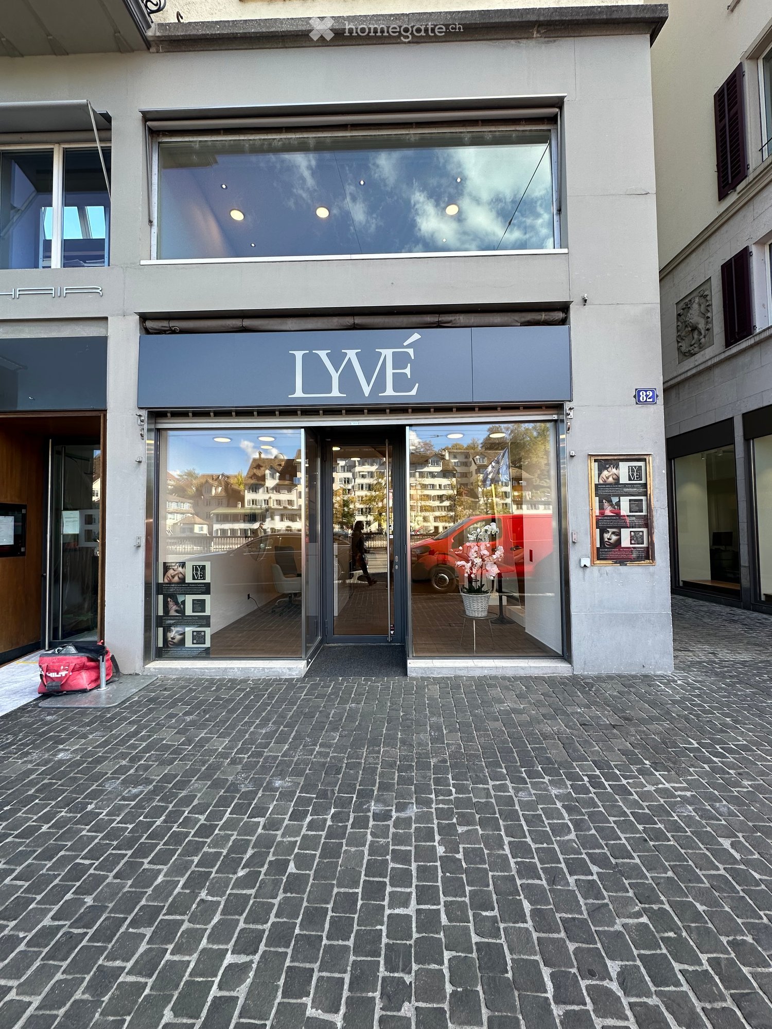 Storefront with large glass windows and a blue awning displaying the sign "LYVE". The entrance features a glass door, and the interior is visible with a few pieces of furniture and a vase with flowers.