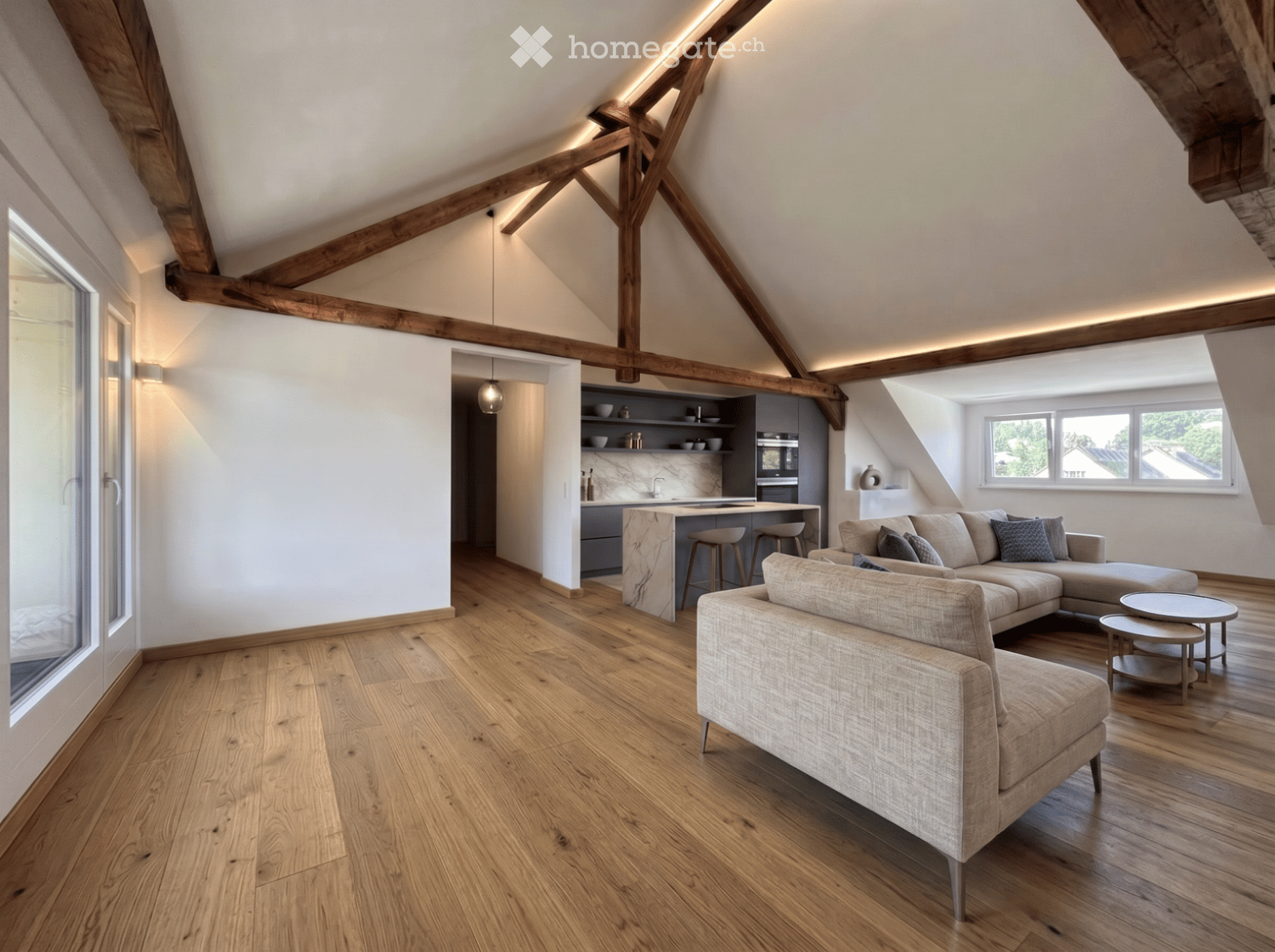 Open-plan living and kitchen area with exposed wooden beams, hardwood flooring, a sectional sofa, coffee table, and a modern kitchen island with stools.