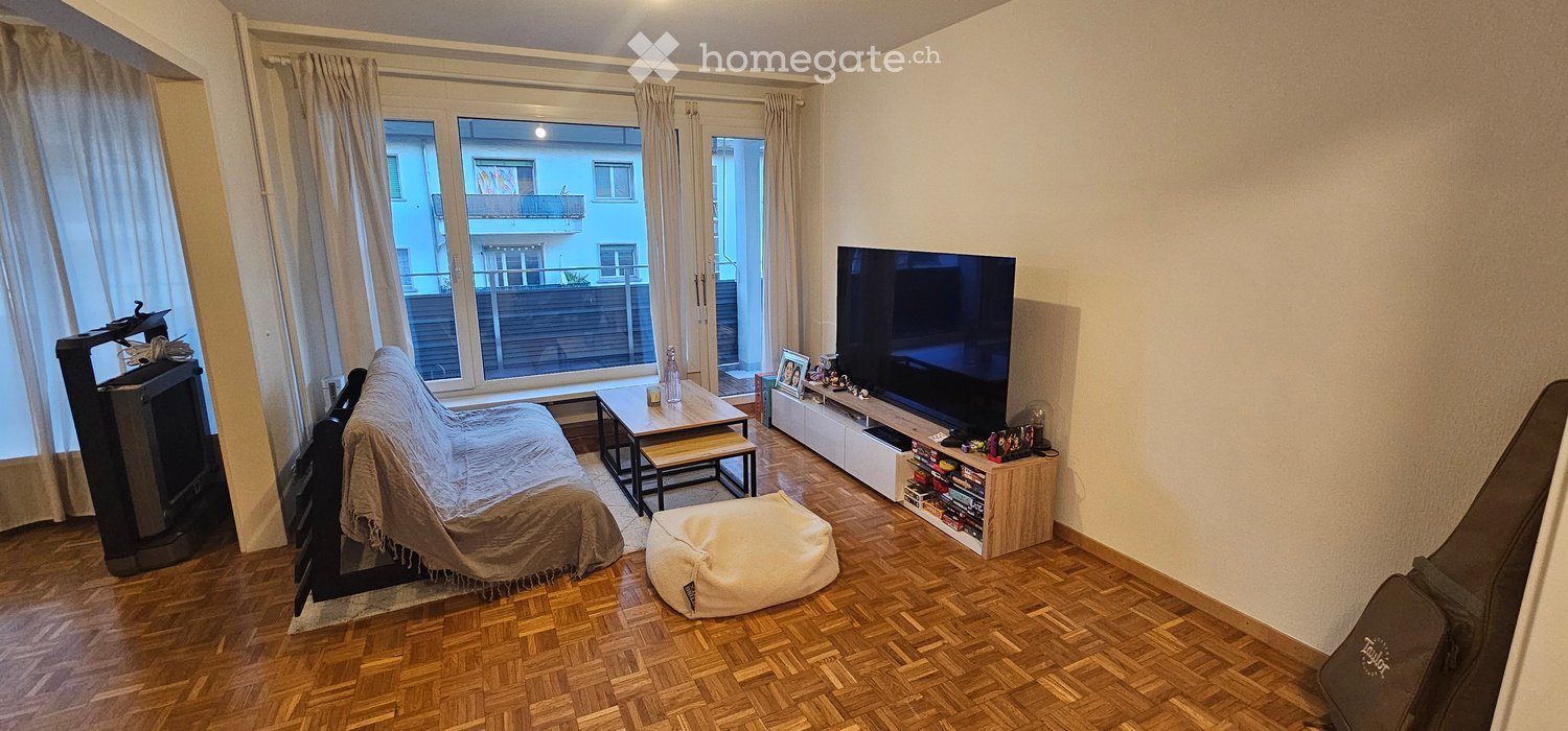 Living room with wooden floor, large window with view, sofa, coffee table, beige pouf, TV stand, TV, and curtain.