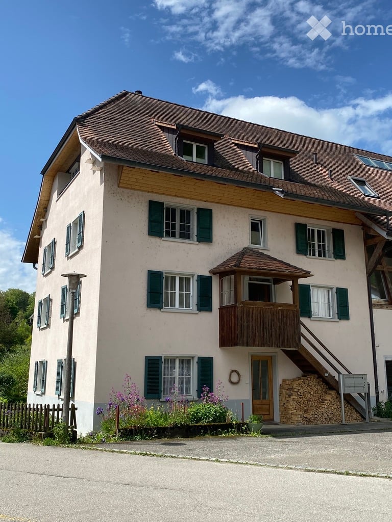Two-story house with a tiled roof, green shutters, and a wooden balcony. The house has a light-colored facade and is surrounded by a small garden with flowers and a stack of firewood.