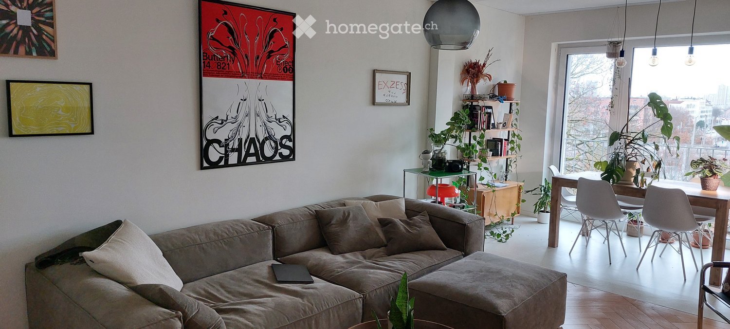 Modern living room with a grey sectional sofa, ottoman, dining table, chairs, shelves, and plants. Large windows offer a city view.