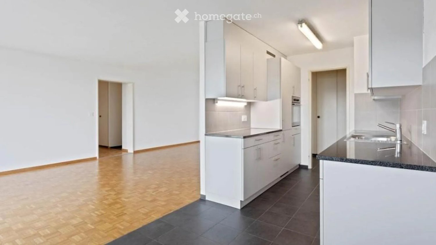 Modern kitchen with white cabinets, dark countertops, and a stainless steel sink. The space features wooden flooring and a clean, minimalist design.