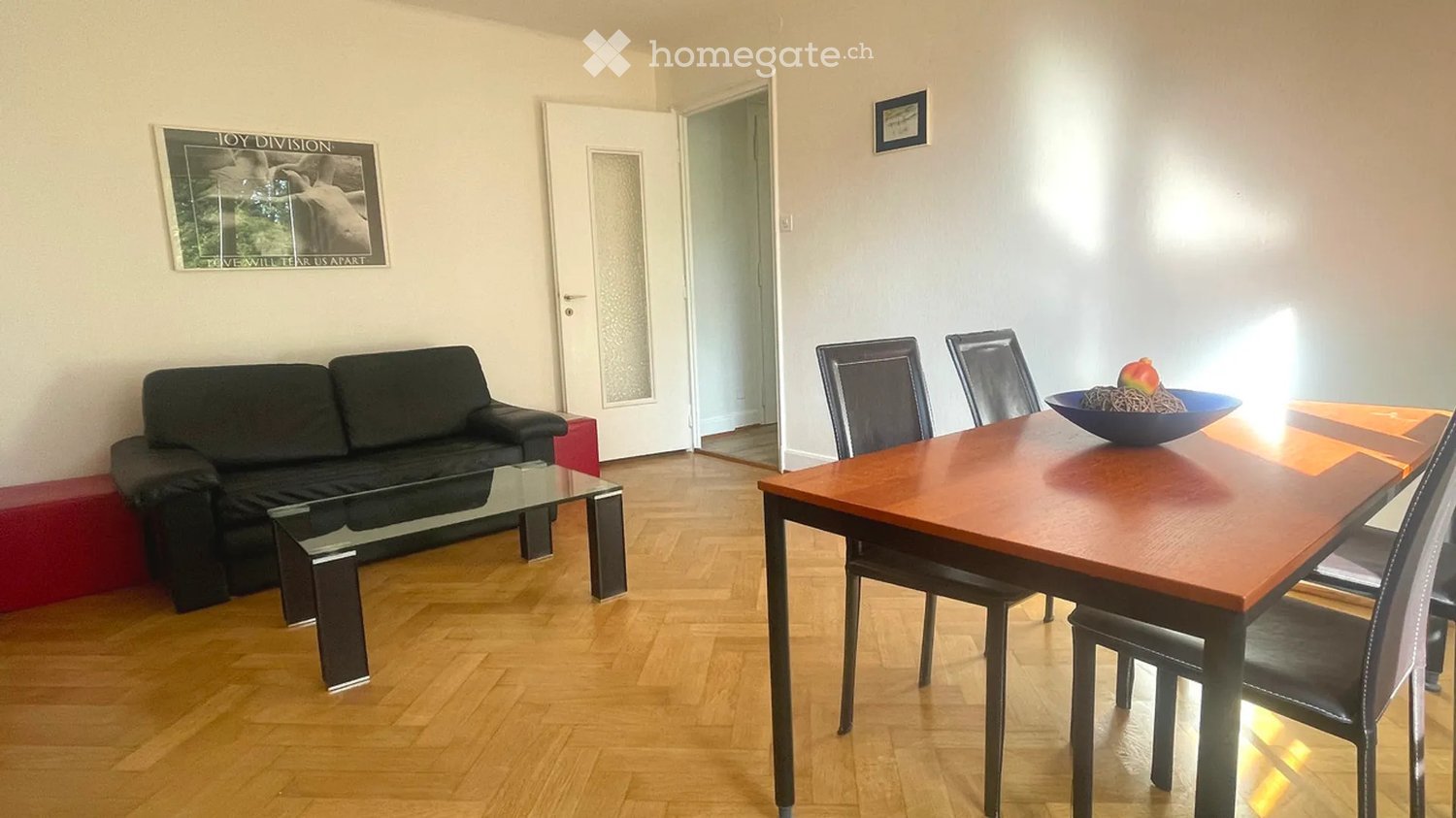 A modern living and dining area with a black sofa, glass coffee table, wooden dining table, and four chairs. A bowl with fruit sits on the dining table. The room features light-colored walls, wooden flooring, and a framed poster on the wall.