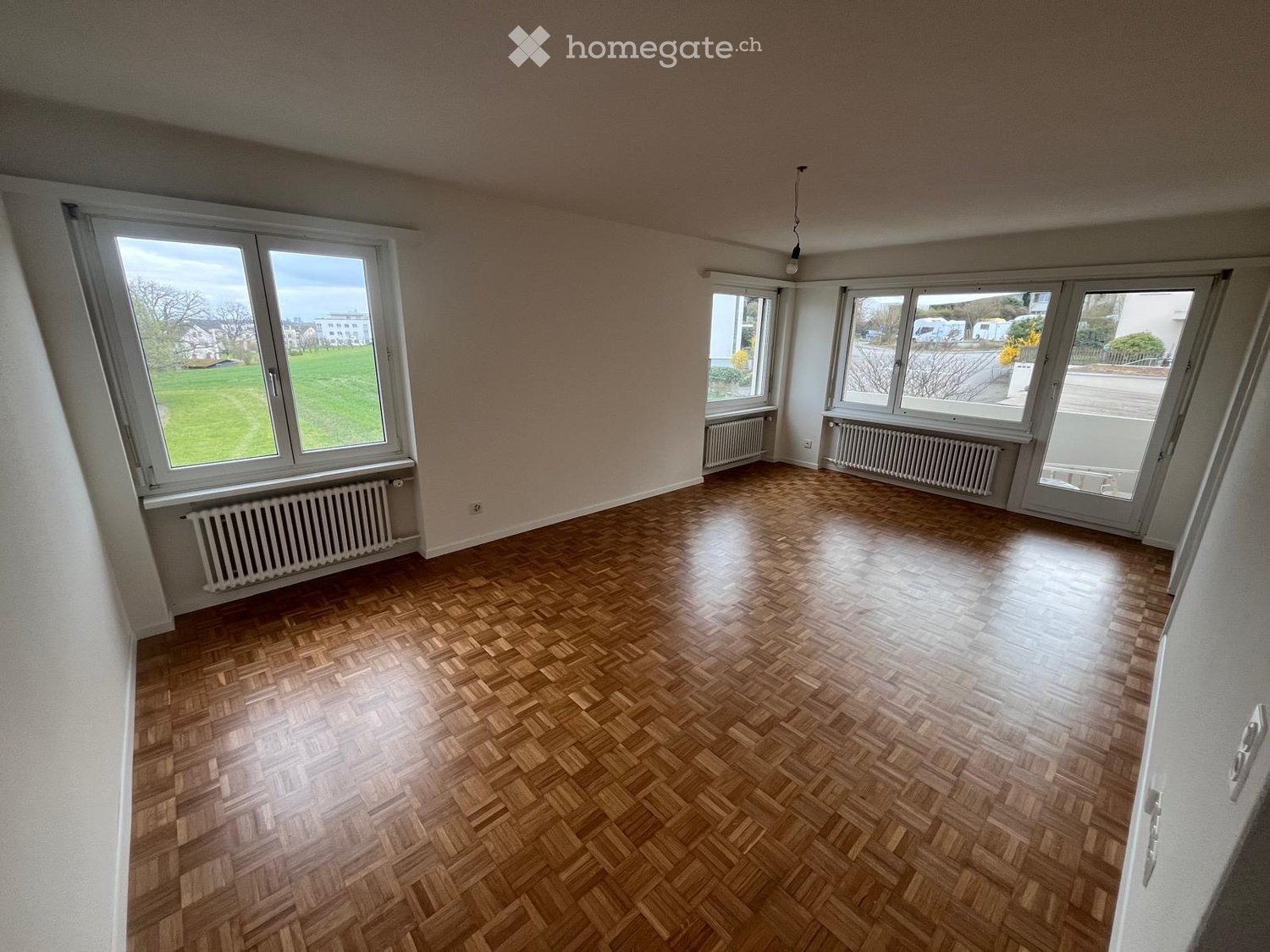 Empty room with wooden herringbone flooring, large windows, radiators, and a view of a green field outside.