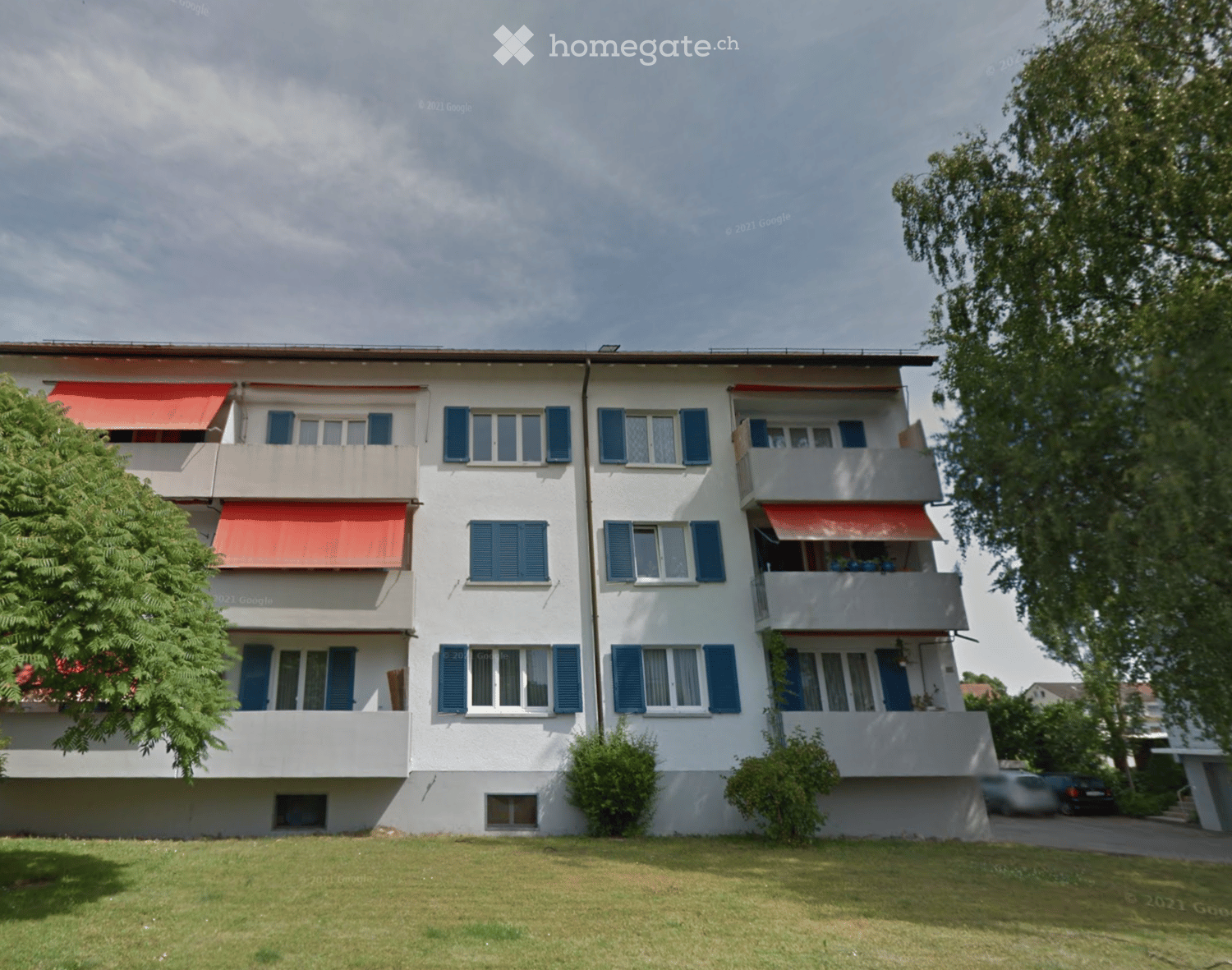 A three-story residential building with white walls, blue shutters, and red awnings over the balconies. The building has multiple windows and is surrounded by greenery and a grassy lawn.