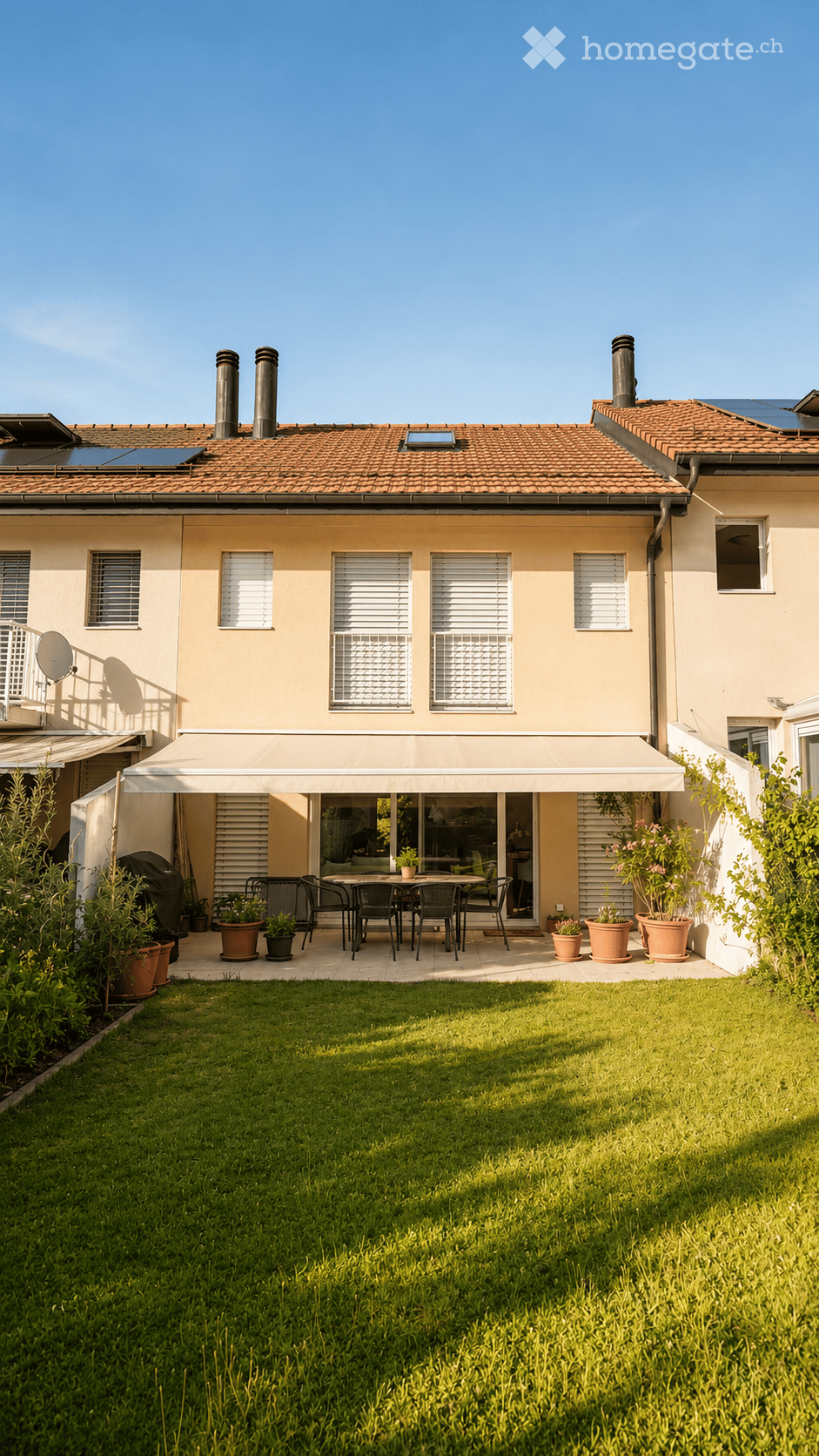 A well-maintained garden with a neatly trimmed lawn, a patio area with a dining set, and potted plants. The house features a tiled roof, multiple windows, and a covered outdoor seating area.