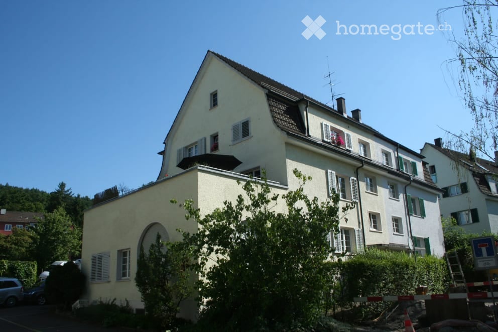 Two-story residential house with light-colored walls, multiple windows, and a small balcony on the upper floor. The house is surrounded by greenery and trees.