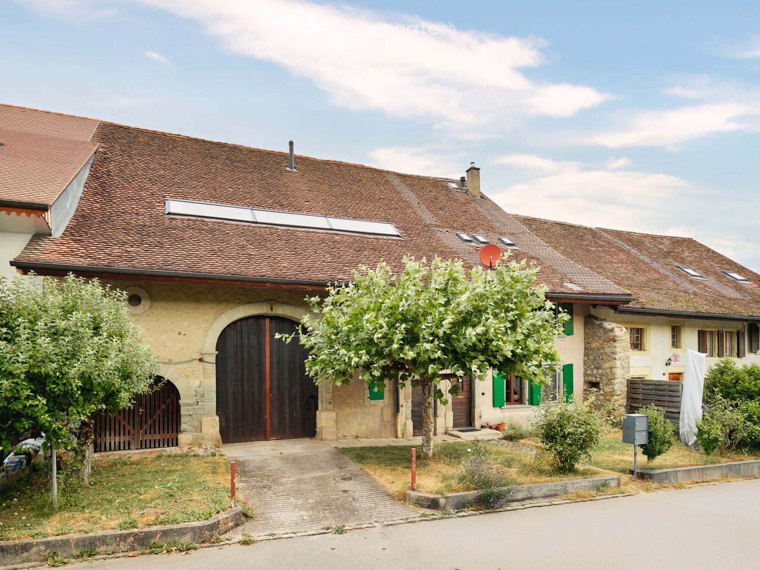 Two-story house with a tiled roof, stone and plaster exterior, large wooden garage door, green shutters, and surrounding greenery.