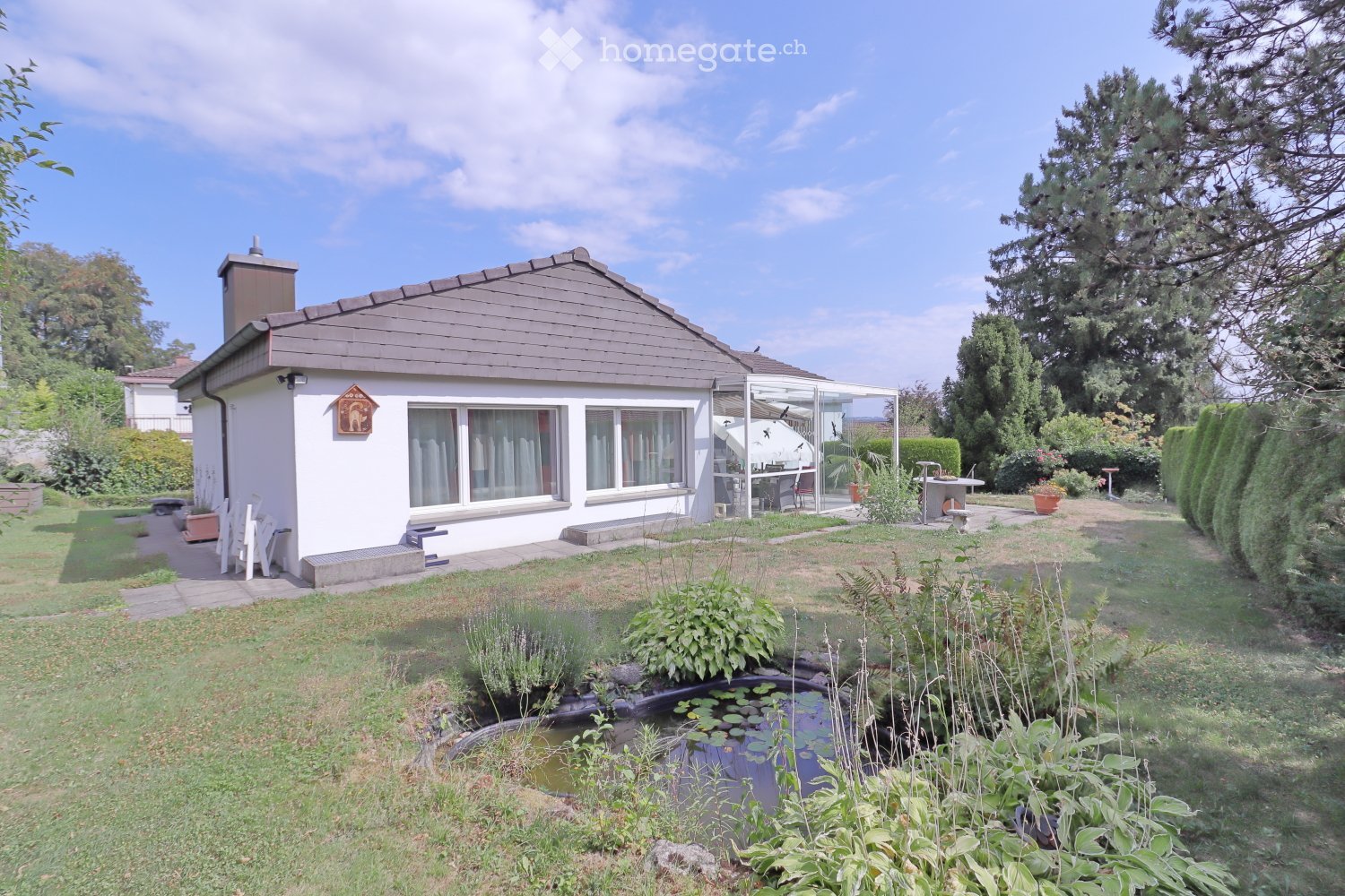 Single-story house with a grey roof and white walls, surrounded by a well-maintained garden with a pond, various plants, and outdoor seating area.