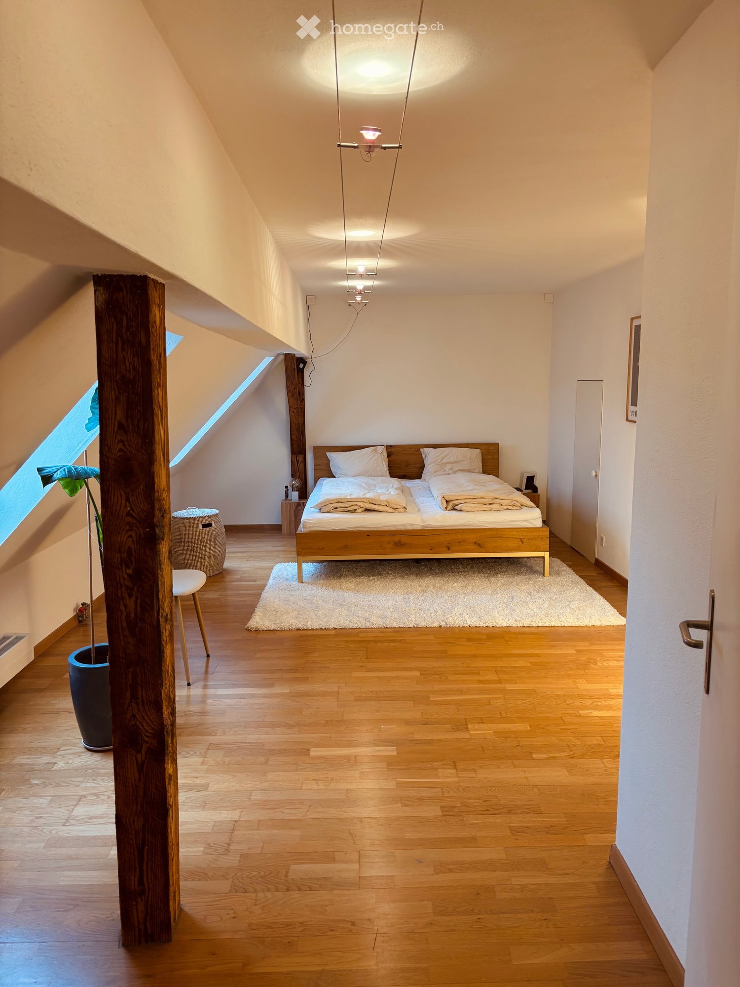 Bright, modern bedroom with wooden floors, large windows, a bed with white bedding, a nightstand, a small stool, a white chair, and a potted plant near the window.