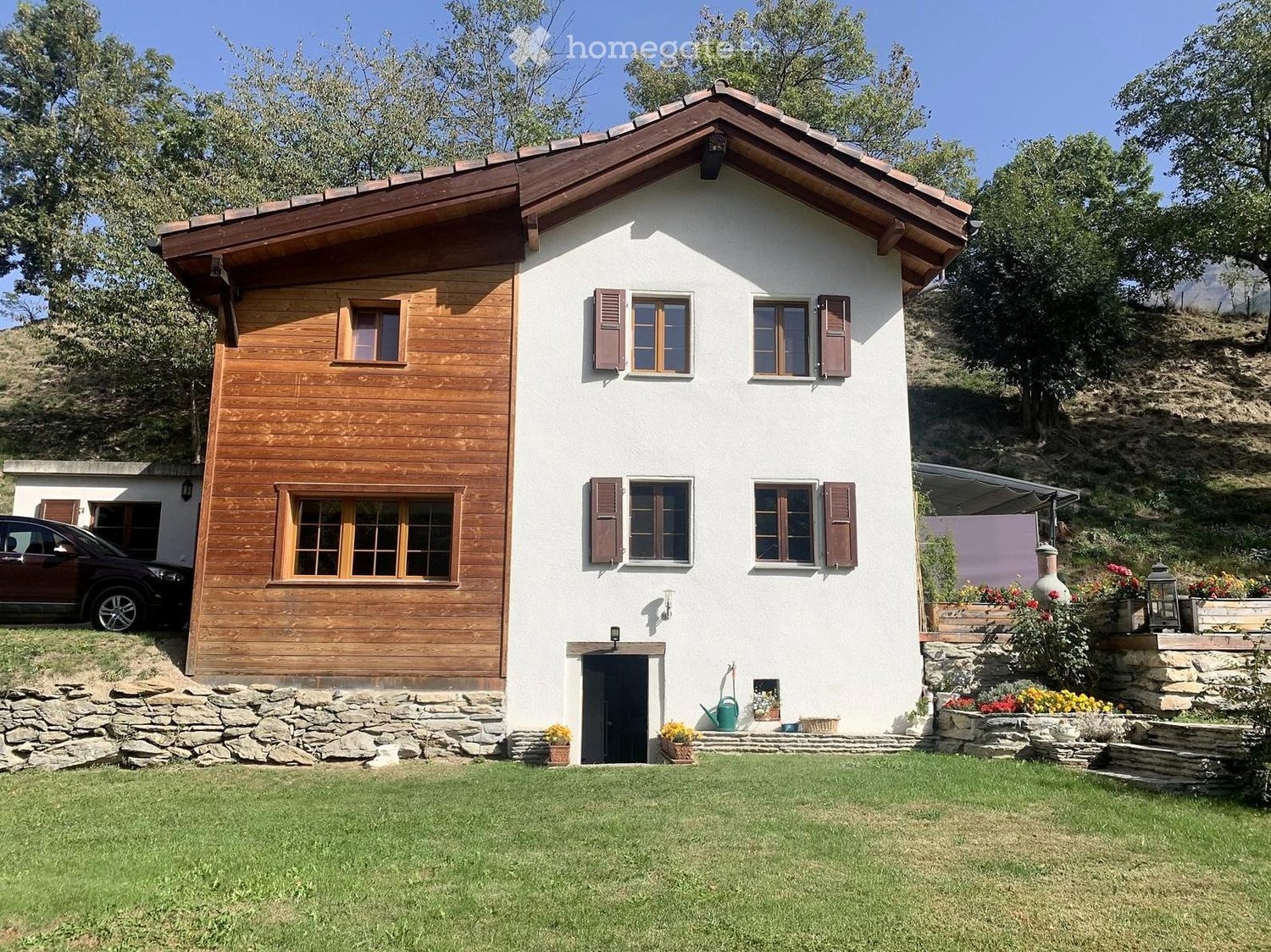 Two-story house with white walls, wooden accents, and a brown tiled roof. Features multiple windows with shutters, a stone foundation, and a well-maintained lawn with flower pots and a parked car nearby.
